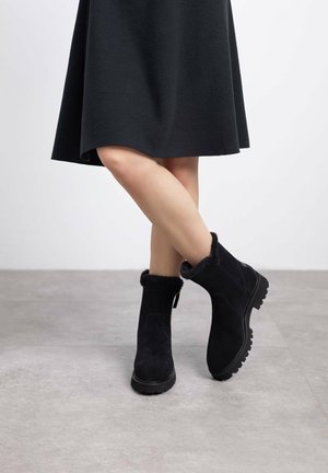 Botas de tobillo de ante negro con un cuello de piel sintética, suela de goma texturizada y cremallera lateral, combinadas con una falda negra.