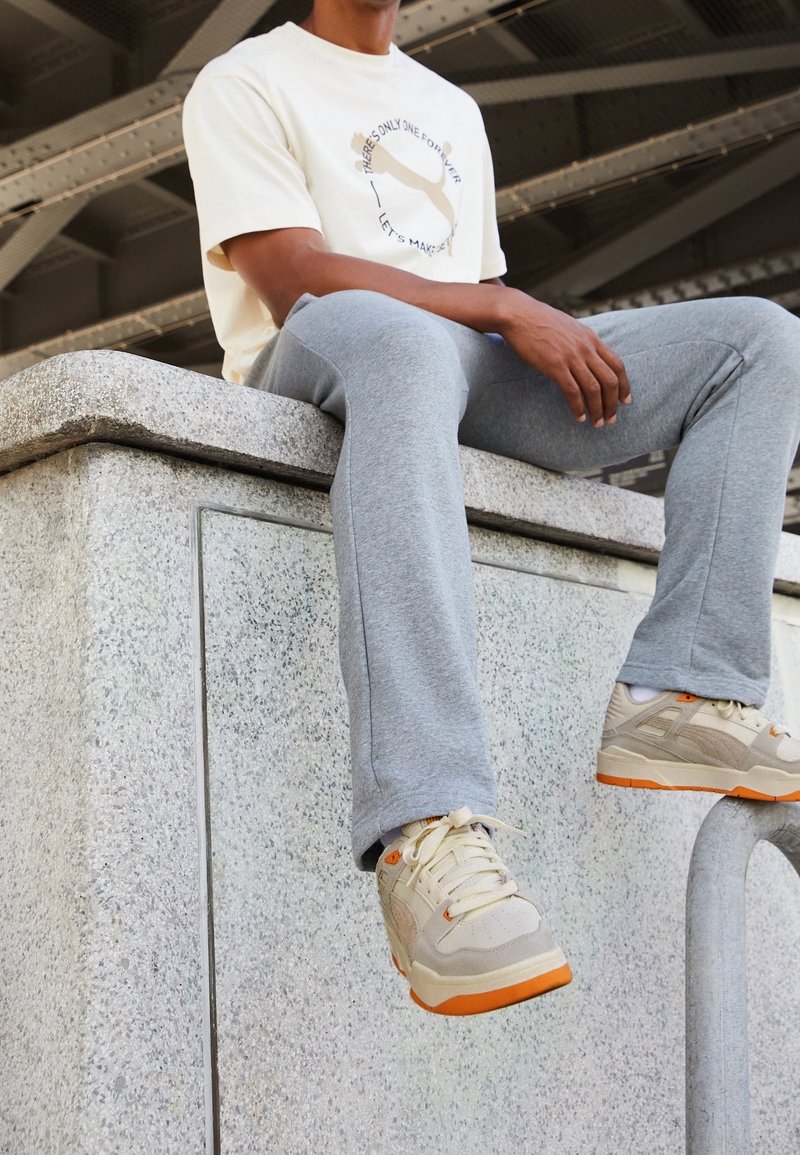 Zapatillas beige con acentos naranjas, pantalones de chándal grises y una camiseta crema con impresión gráfica, sentado en un muro de piedra.