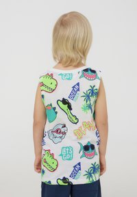 Maglietta bianca senza maniche con stampati colorati di cartoni animati raffiguranti dinosauri, squali e palme, accompagnati da elementi testuali in verde brillante e blu.
