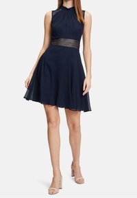 Marinblå ärmlös klänning med en plisserad bodice och spetsdetalj i midjan, med en utsvängd kjol i lätt material, parat med ljusa klackar.