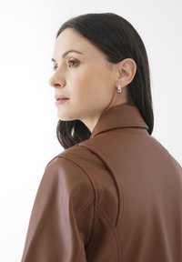 Brun läderjacka med hög krage och markerade axelsömmar. Silverfärgade stiftörhängen framhäver modellens profil mot en ljus bakgrund.