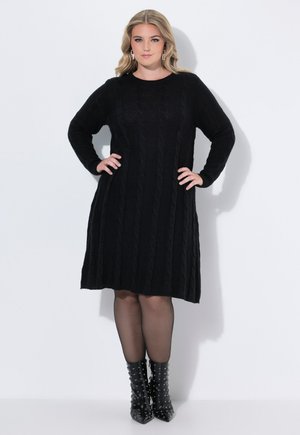 Vestido suéter de punto trenzado negro con mangas largas, escote redondo y dobladillo acampanado. Combinado con botas de tobillo negras brillantes con tachuelas.