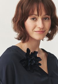 Tengerészkék blúz V-nyakkivágással, amelyen egy nagy fekete szövetvirág díszítés található a bal oldalon. Arany lánc nyaklánc látható.