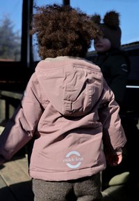 Veste imperméable rose avec capuche et poignets élastiques, arborant un logo blanc mikk-line dans le dos. Texture confortable avec doublure douce.