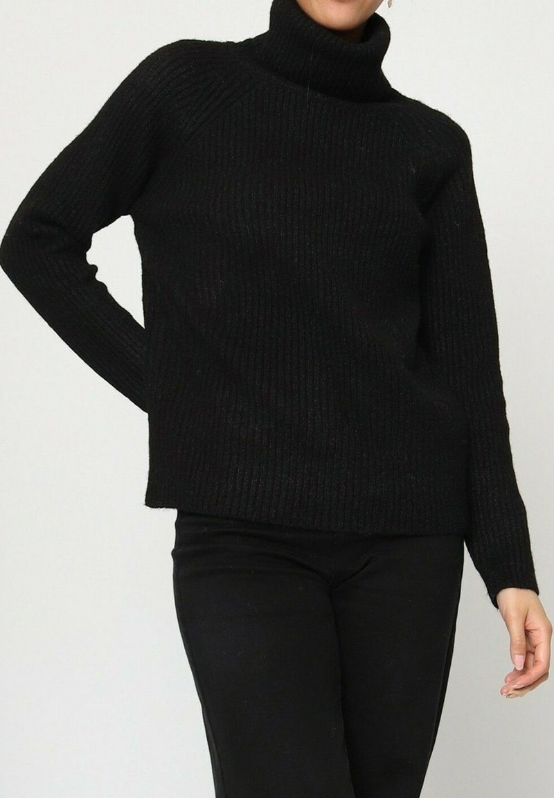 Personne portant un pull col roulé noir à côtes et un pantalon noir, posant avec une main sur la hanche devant un fond uni.