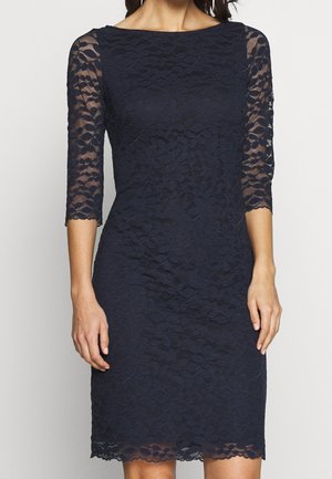 Woman wearing a knee-length, navy blue lace dress with three-quarter sheer sleeves and a scalloped hem.