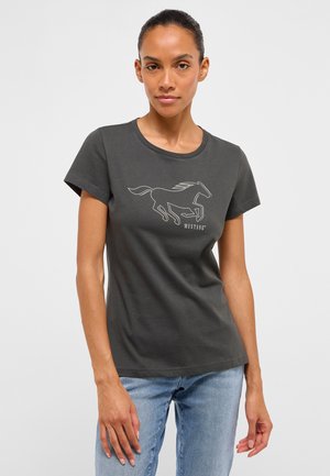 Frau trägt ein dunkelgraues T-Shirt mit einer weißen Umrisszeichnung eines Mustang-Pferdes und hellblauen Jeans vor einem schlichten weißen Hintergrund.