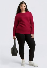 Bordeauxrode gebreide trui met structuur en een ronde hals, gecombineerd met zwarte taps toelopende broek en witte sneakers, met een kleine zwarte handtas.