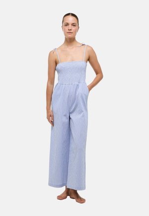 Femme debout pieds nus portant une combinaison bleu clair sans manches à jambes larges avec un corsage froncé et des bretelles nouées aux épaules sur un fond uni.
