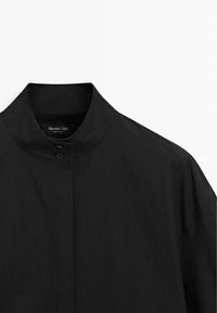 Schwarze Jacke mit hohem Kragen, Knopfdetail und glattem, strukturiertem Stoff. Einfaches Design, ohne Muster oder sichtbare Hardware-Akzente.