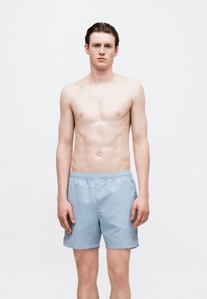Young man with short brown hair wearing light blue swim shorts, standing against a plain white background, facing forward with a neutral expression.