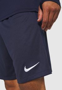 Mężczyzna w granatowych szortach sportowych Nike z białym logo swoosh na lewej udzie, na jednolitym tle.