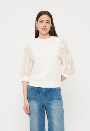 Vrouw met lang bruin haar, gekleed in een wit bovenstuk met kanten pofmouwen en blauwe spijkerbroek, staand tegen een effen witte achtergrond.