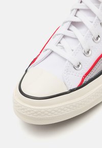 Hvite lerrets sneakers med røde og svarte detaljer, gummitåhette og teksturert hvit såle. Har metall øyesplitter og hvite snørebånd.