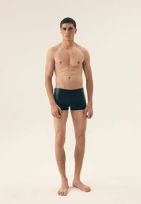 Czarne kąpielówki z niebieskimi akcentami, dopasowany krój, gładka faktura i krótka długość, noszone przez męskiego modela na neutralnym tle.