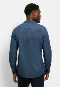 Mann mit kurzen, lockigen Haaren, der ein langärmliges, blaues Hemd trägt, von hinten vor einem schlichten weißen Hintergrund gezeigt.