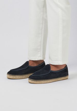Pieds portant des espadrilles en daim noir avec semelles en jute, associés à un pantalon blanc retroussé sur un fond clair uni.