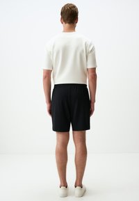 Chemise blanche à manches courtes, shorts noirs avec taille élastique. Modèle portant des baskets blanches, vu de dos sur un fond uni.