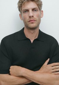 Svart kortärmad polo skjorta med texturerad tyg, med krage och v-ringning, bärs av en man med armarna i kors.