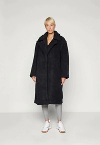 Person with short blonde hair wearing a long black teddy coat, gray leggings, and white sneakers standing on a plain background.