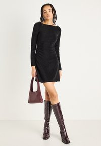 Abito nero con maniche lunghe, dal design aderente, in tessuto testurizzato, abbinato a stivali al ginocchio bordeaux in pelle imitazione coccodrillo e una borsa coordinata.