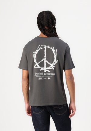 Personne avec des cheveux tressés portant un t-shirt gris foncé avec un symbole de paix blanc fait de bâtons et du texte au dos.