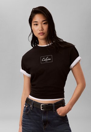 Młoda kobieta z długimi ciemnymi włosami, ubrana w czarny krótki t-shirt z białym obszyciem oraz ciemne dżinsy z czarnym paskiem, ręka w kieszeni.