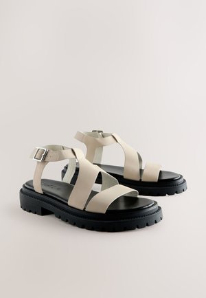 Beige leather sandals with cross straps and a buckle detail. Textured black rubber sole with a thick, treaded design.