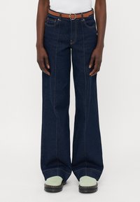 Temno modri široki jeans z visokim pasom, ki imajo kontrastne šive in rjav pas s krožnim sponko. Črne platforme.