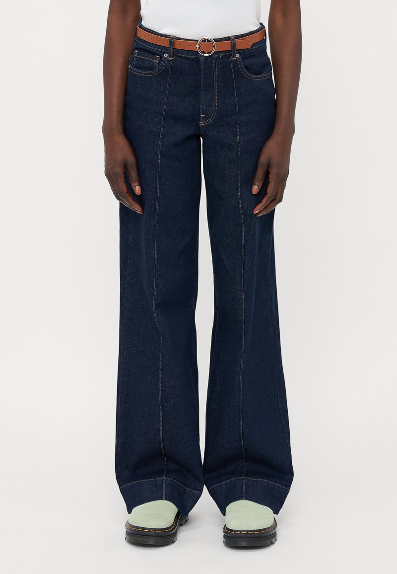 Temno modri široki jeans z visokim pasom, ki imajo kontrastne šive in rjav pas s krožnim sponko. Črne platforme.