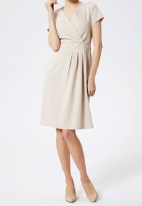 Vestido envolvente de color beige con mangas cortas, falda plisada y un corsé ajustado. Tela suave y diseño a la altura de la rodilla. Combinado con tacones color nude.