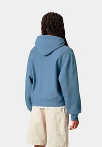Sudadera con capucha de color azul claro, de corte relajado, con puños y dobladillo acanalados. La vista posterior muestra un diseño sencillo y una tela suave y texturada.