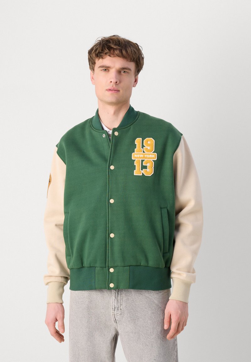 Veste varsity vert et crème avec broderies "19" et "13 NEW YORK", boutons-pression, et poignets et ourlet côtelés.