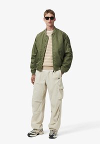 Bomber jacket verde oliva con polsini a coste, maglione a righe con perline e pantaloni cargo crema. Occhiali da sole neri e scarpe sportive completano il look.