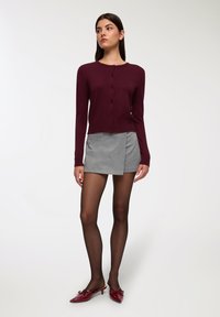 Cardigan bordeaux con scollo rotondo e chiusura a bottoni, abbinato a una gonna a portafoglio grigia. Modella che indossa collant neri e mule con tacco rossi.