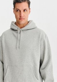 Jasnoszara bluza z kapturem, wykonana z miękkiego materiału, z dużą przednią kieszenią, sznurkami w kapturze i luźnym krojem.