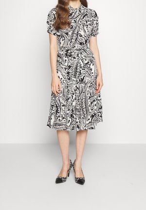 Femme portant une robe midi à manches courtes à motif cachemire noir et blanc, ceinturée à la taille, et des escarpins noirs à motifs.