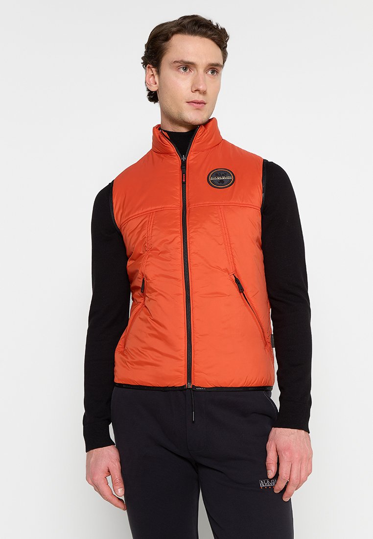 Pomarańczowa kamizelka izolacyjna z wysokim kołnierzem, zapinana na zamek z przodu, z kieszeniami po bokach i okrągłą naszywką logo na klatce piersiowej. Gładka, puszysta faktura.