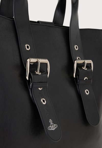 Κοντινή λήψη μαύρων δερμάτινων ιμάντων τσάντας με ασημένιες αγκράφες και μεταλλικά μάτια, με ένα μικρό ανάγλυφο λογότυπο κοντά στο άκρο του ιμάντα.
