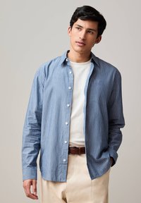 Giovane uomo con capelli neri che indossa una camicia aperta a righe blu sopra una t-shirt bianca e pantaloni beige, in piedi con una mano in tasca.