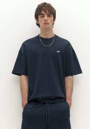 Junger Mann trägt dunkel übergroßes T-Shirt und passende Hose, steht vor weißem Hintergrund mit den Händen in den Taschen.