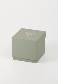 Caja cuadrada texturizada en gris-verde de Guess con logotipo triangular dorado en la tapa, contra un fondo blanco.