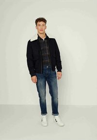 Casaco bomber preto com colarinho de shearling branco, combinado com uma camisa de botão xadrez, jeans azuis e ténis brancos com detalhes pretos.
