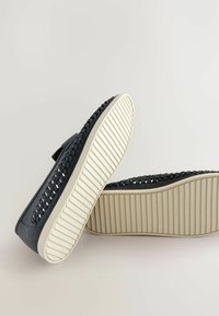 Zapatos de lona gris oscuro tipo slip-on con acentos trenzados, suela de goma blanca y fondo texturizado; diseñados para uso casual con una estructura transpirable.