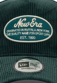 Grønn cordfløyelslue med "New Era"-logo i hvitt og turkis, med justerbar stropp og et broderi-patch med tekstdetaljer.