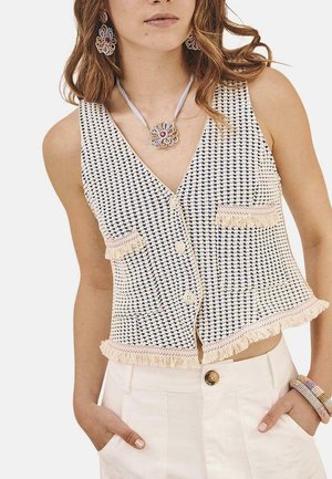 Chaleco corto blanco y marino con textura tejida, escote en V, dos bolsillos y detalle de dobladillo deshilachado. Combinado con pantalones blancos de tiro alto.