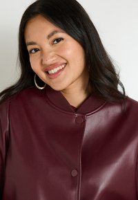 Chaqueta de cuero sintético burdeos con cuello redondo, cierre de botón a presión y acentos acanalados. Textura suave con un acabado liso.