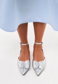 Scarpe ballerine argentate a punta, con un raffinato fiocco testurizzato, cinturini alla caviglia e finitura metallica lucida, abbinati a un vestito azzurro chiaro.