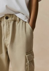 Pantalons cargo beiges avec une finition texturée, ceinture élastique, poches latérales et détails de surpiqûres visibles, associés à un t-shirt blanc à col rond.