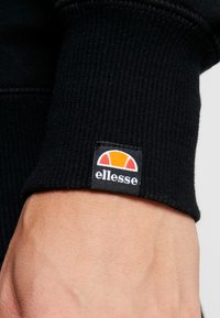 Schwarzer gerippter Sweatshirtärmel mit einem sichtbaren Patch, der ein buntes Logo in Orange, Weiß und Schwarz zeigt. Strukturiertes Material und Nahtdetails.